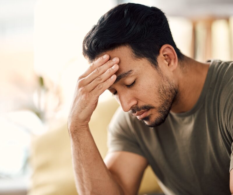 A man in a green shirt sits with his hand on his forehead, looking distressed. - Headache