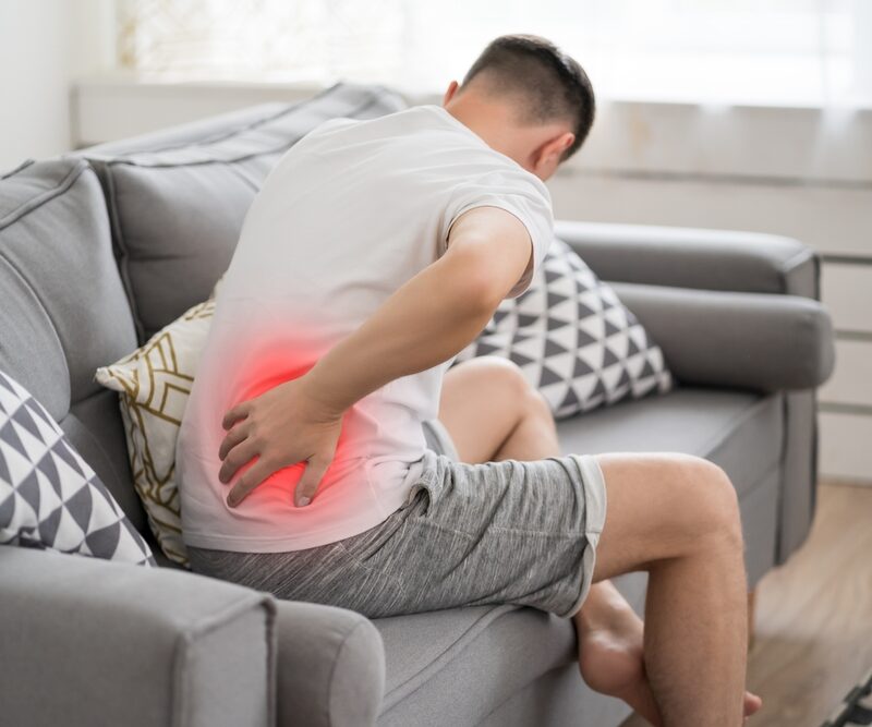 A man sitting on a couch holds his lower back in pain, with a red glow indicating discomfort. - Sciatica